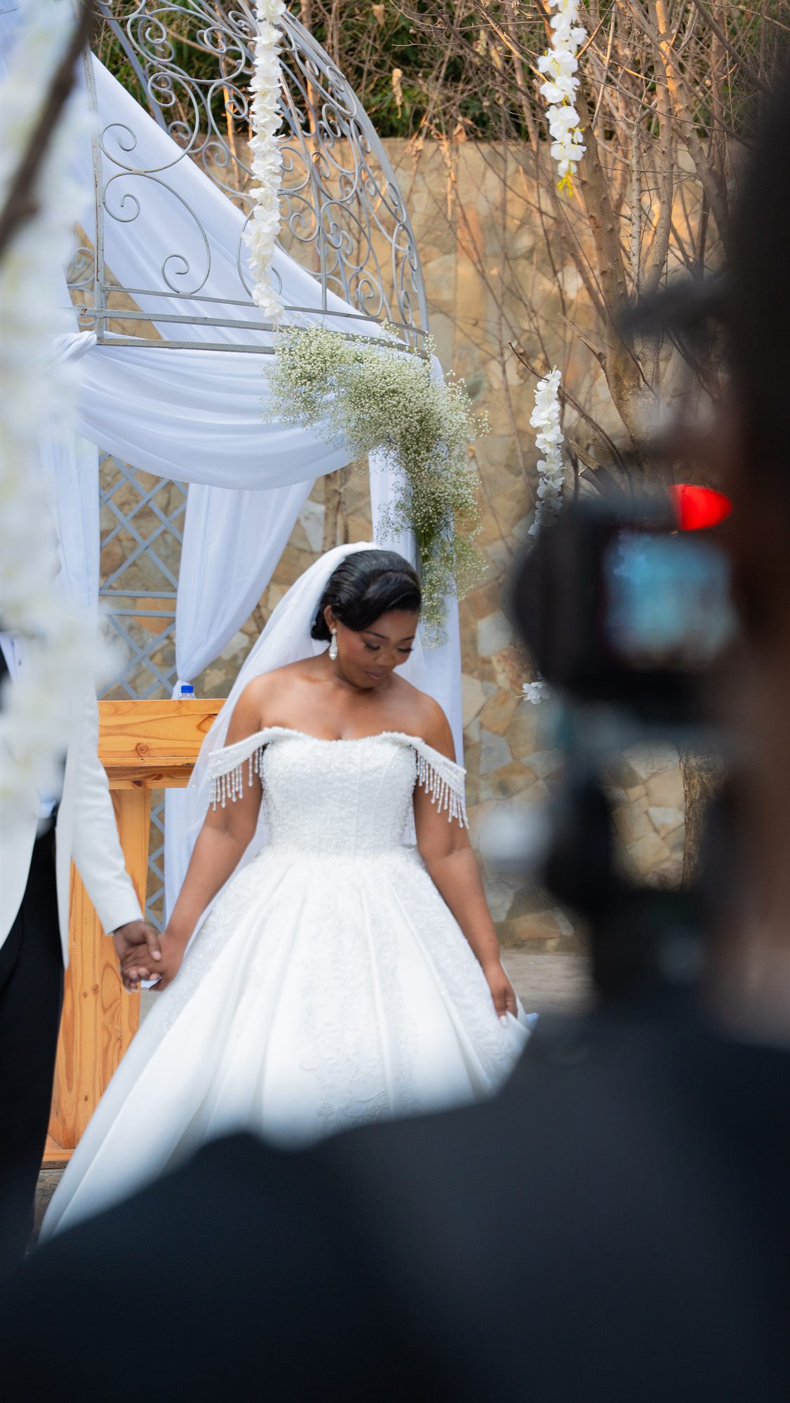 Bride walking toward the ceremony arch, seen past a camera operator